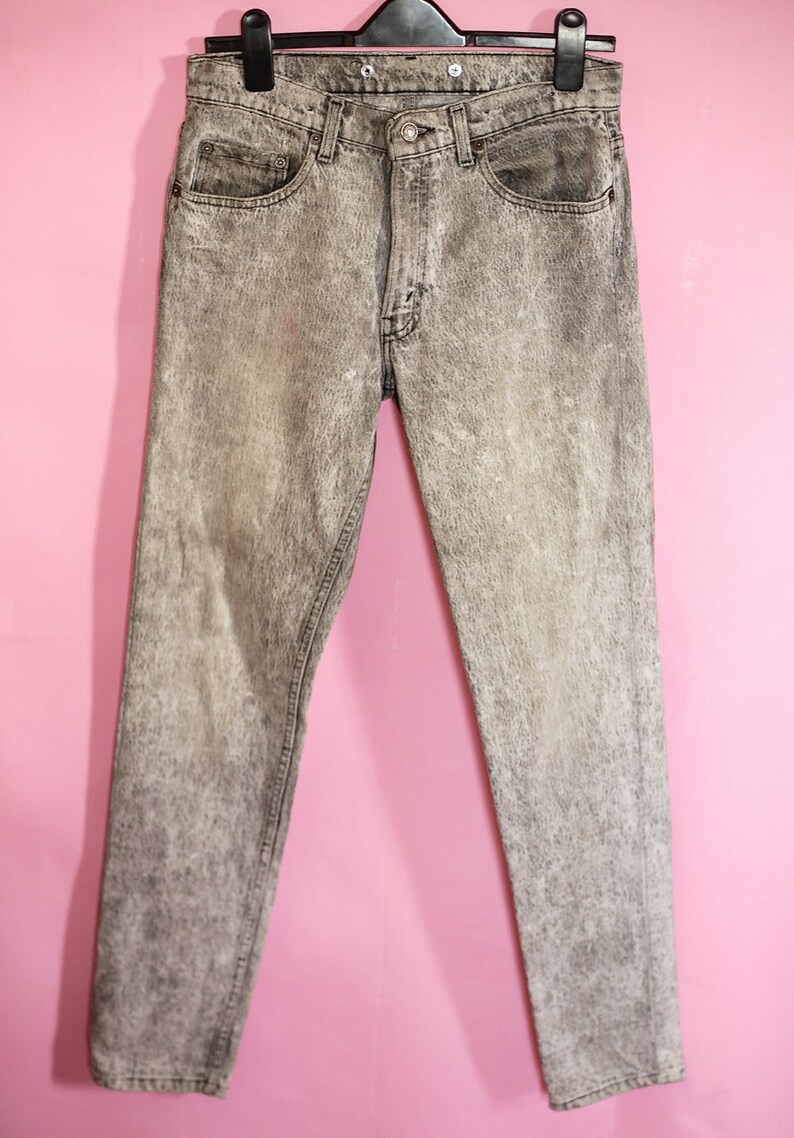 May include: Grey acid wash jeans with a straight leg fit. The jeans are hanging on a black hanger against a pink background.