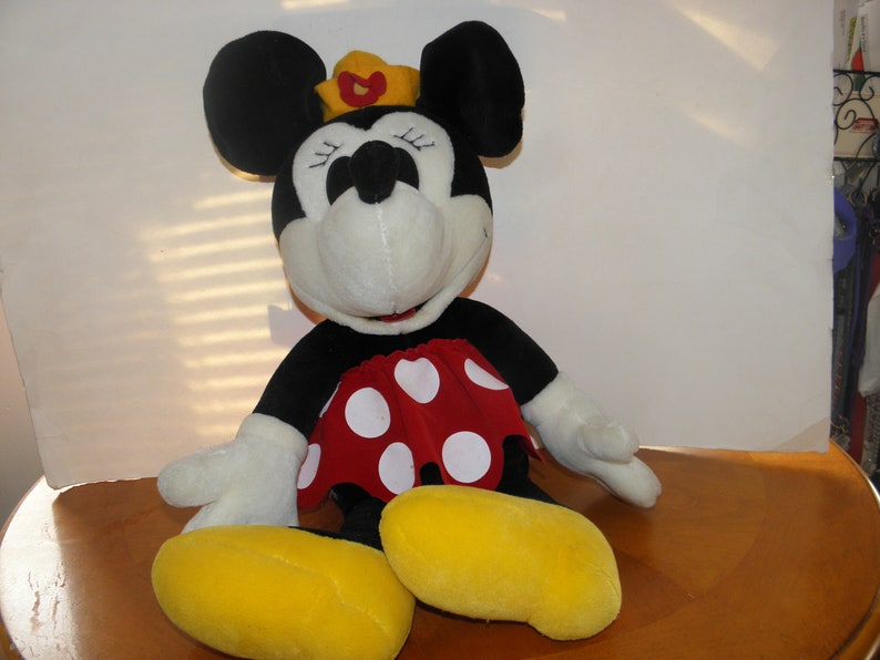 May include: A large plush Minnie Mouse toy wearing a red and white polka dot dress and a gold crown. She has black ears and a white face with black eyes and a black nose.