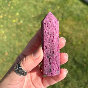 May include: A pink ruby crystal obelisk with a textured surface. The crystal is held in a hand against a blurred green background. The obelisk is a rectangular shape with a pointed top.