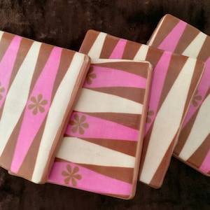 May include: Set of four square coasters with a retro design. Each coaster features a pattern of diagonal stripes in pink, white, and brown, with small flower accents. The coasters are arranged on a brown surface.