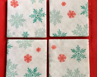 May include: Set of four white ceramic coasters with a snowflake pattern in teal and red. The coasters are square and have a smooth, glossy finish.