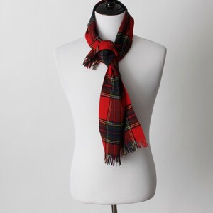 May include: A red and black plaid scarf with fringed edges is draped around a white mannequin. The scarf features a classic tartan pattern with green and yellow accents. The scarf is a winter accessory.