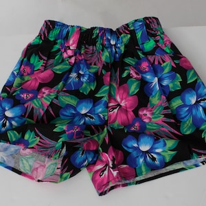 Puede incluir: Pantalones cortos negros con cintura elástica, con un estampado floral vibrante. El diseño incluye grandes flores de hibisco azules y rosas, hojas verdes y detalles en rosa. Los pantalones cortos están hechos de tela ligera.