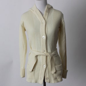 Könnte beinhalten: Ein cremefarbener Strickcardigan mit Kapuze, langen Ärmeln und Knopfleiste. Er hat einen Gürtel und Taschen.