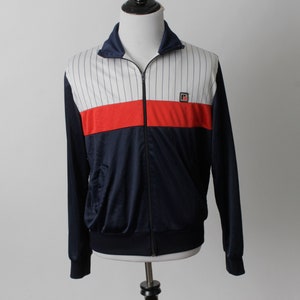 May include: A navy blue track jacket with a white and red color block design. The jacket has a full zipper, a stand-up collar, and a small logo on the chest. The white section has vertical black pinstripes.
