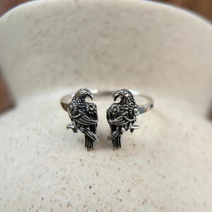 Op de afbeelding: Een zilveren ring met twee gedetailleerde ravenfiguren die elkaar aankijken. De ring is open en verstelbaar.