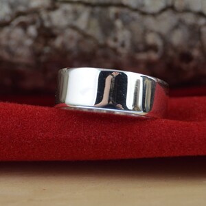 May include: A polished silver ring with a flat top and sides. The ring is set against a red background, reflecting light. The ring's design is simple and modern, suitable for everyday wear. The ring is approximately 0.3 inches wide.