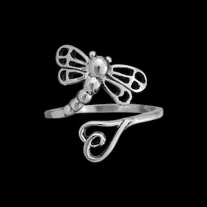 May include: A silver ring with a dragonfly motif. The dragonfly has intricate wing detailing and a segmented body. The ring band curves into a heart shape. The ring is displayed against a black backdrop.