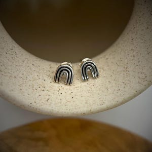 May include: A pair of silver rainbow stud earrings. The earrings are small and delicate, and they feature a simple, minimalist design.