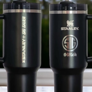 May include: Two black stainless steel travel mugs with handles and lids. The mug on the left has the text 'STANLEY + 30oz SAUER' engraved on the side. The mug on the right has the text 'STANLEY' and a logo engraved on the side. Both mugs have a straw in them.
