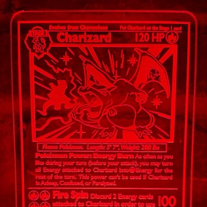 Könnte beinhalten: Ein rotes, beleuchtetes Charizard Pokémon-Karten-Nachtlicht. Die Karte zeigt eine detaillierte Illustration von Charizard, mit Text wie "120 HP" und "Fire Spin". Das Nachtlicht ist an einer schwarzen Basis angeschlossen.