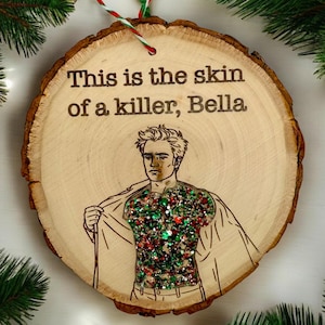 May include: A circular wooden Christmas ornament with the text "This is the skin of a killer, Bella." It features an illustration of a person with a glittery torso. The ornament is suspended by a red, green, and white string.