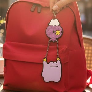 May include: A red backpack with a matching keychain featuring two cartoon characters. The keychain is purple, white, and yellow. The backpack is sitting on a wooden table outside, with a cup of coffee and a phone.