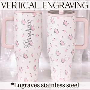 May include: Two stainless steel tumblers with pink handles and lids. The tumblers are white with a floral pattern of pink roses. One tumbler has the name "Seraphena" engraved vertically. Text at the top reads "VERTICAL ENGRAVING" and at the bottom "Engraves stainless steel."