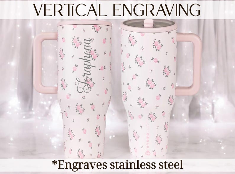 May include: Two stainless steel tumblers with pink handles and lids. The tumblers are white with a floral pattern of pink roses. One tumbler has the name "Seraphena" engraved vertically. Text at the top reads "VERTICAL ENGRAVING" and at the bottom "Engraves stainless steel."
