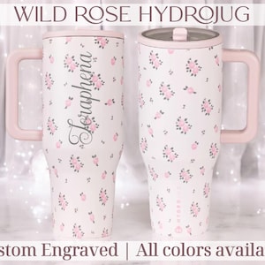 May include: Two white Hydrojugs with pink handles and lids, adorned with pink floral patterns. One jug features the name "Seraphena" in script. The text "WILD ROSE HYDROJUG" and "Custom Engraved | All colors available" are displayed.