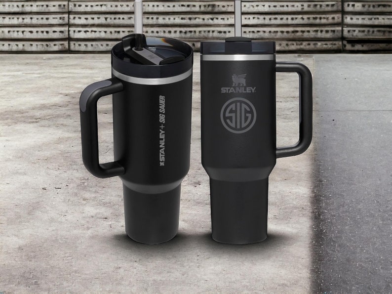 Puede incluir: Dos tazas de viaje de acero inoxidable negro con asas. La taza de la izquierda tiene el texto "#STANLEY + SIG SAUER" en el lateral. La taza de la derecha tiene el texto "STANLEY" y un logotipo de SIG SAUER en el lateral.