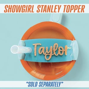 May include: An orange and teal Stanley topper with a straw. The topper has the name "Taylor" in gold glitter lettering. The text "SHOWGIRL STANLEY TOPPER" is at the top. The text "*SOLD SEPARATELY*" is at the bottom.