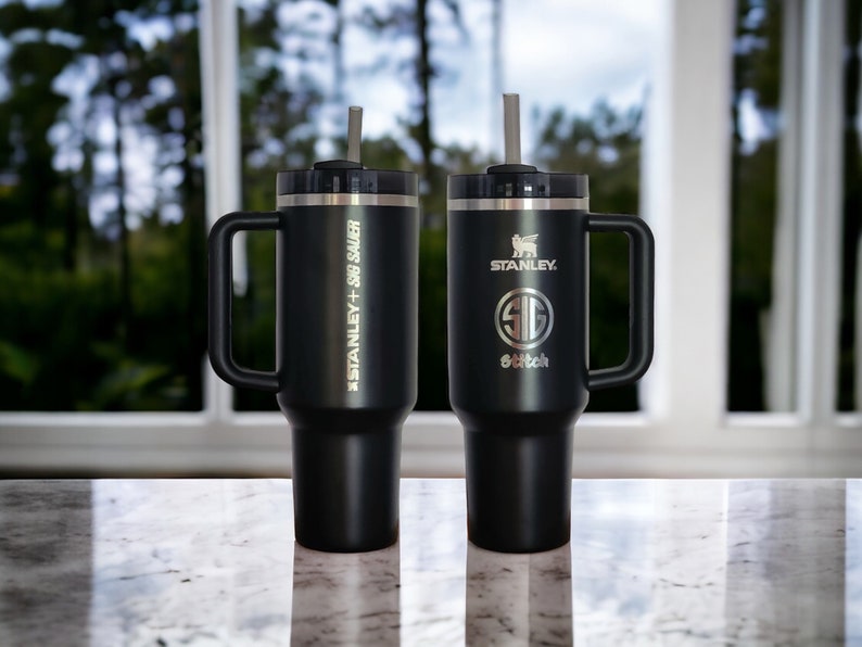 May include: Two black stainless steel travel mugs with handles and lids. The mug on the left has the text 'STANLEY + 30oz SAUER' engraved on the side. The mug on the right has the text 'STANLEY' and a logo engraved on the side. Both mugs have a straw in them.