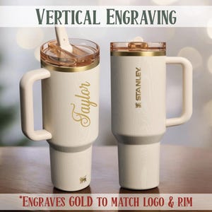 May include: Two white insulated tumblers with gold accents and handles. One tumbler has the name "Taylor" in gold script, the other has the "Stanley" logo. The text "VERTICAL ENGRAVING" and "*ENGRAVES GOLD TO MATCH LOGO & RIM" are also visible.