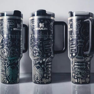 May include: Three black Stanley tumblers with silver detailing. Each tumbler features a handle and a lid. The designs include text and illustrations related to the word "Twilight", including a wolf, a strawberry, and a chess piece.