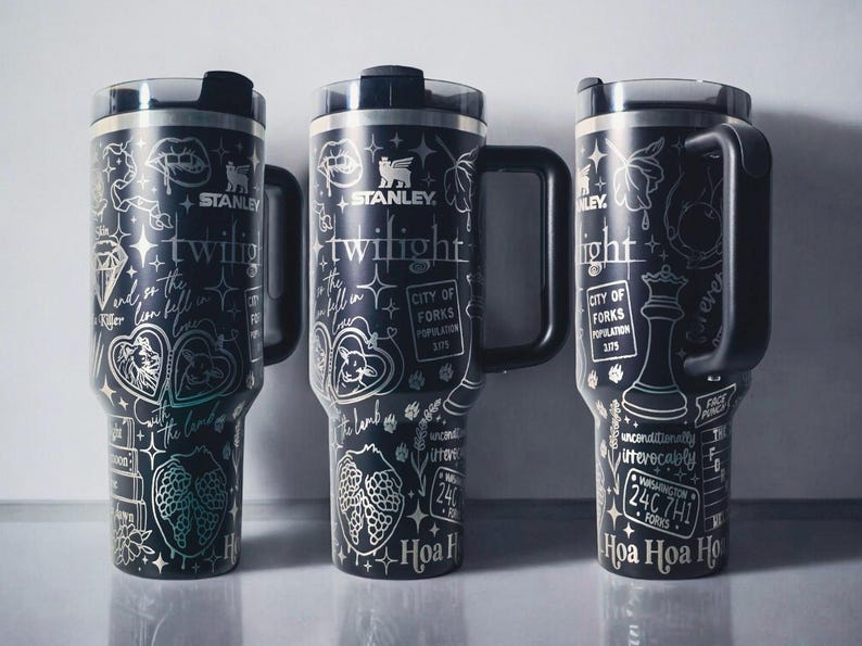 May include: Three black Stanley tumblers with silver detailing. Each tumbler features a handle and a lid. The designs include text and illustrations related to the word "Twilight", including a wolf, a strawberry, and a chess piece.