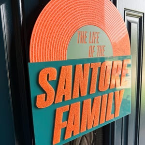 May include: A decorative sign with the text "THE LIFE OF THE SANTORRE FAMILY." The sign features a retro vinyl record design in orange and teal. The text is in a bold, orange font, and the sign is hanging on a teal door.