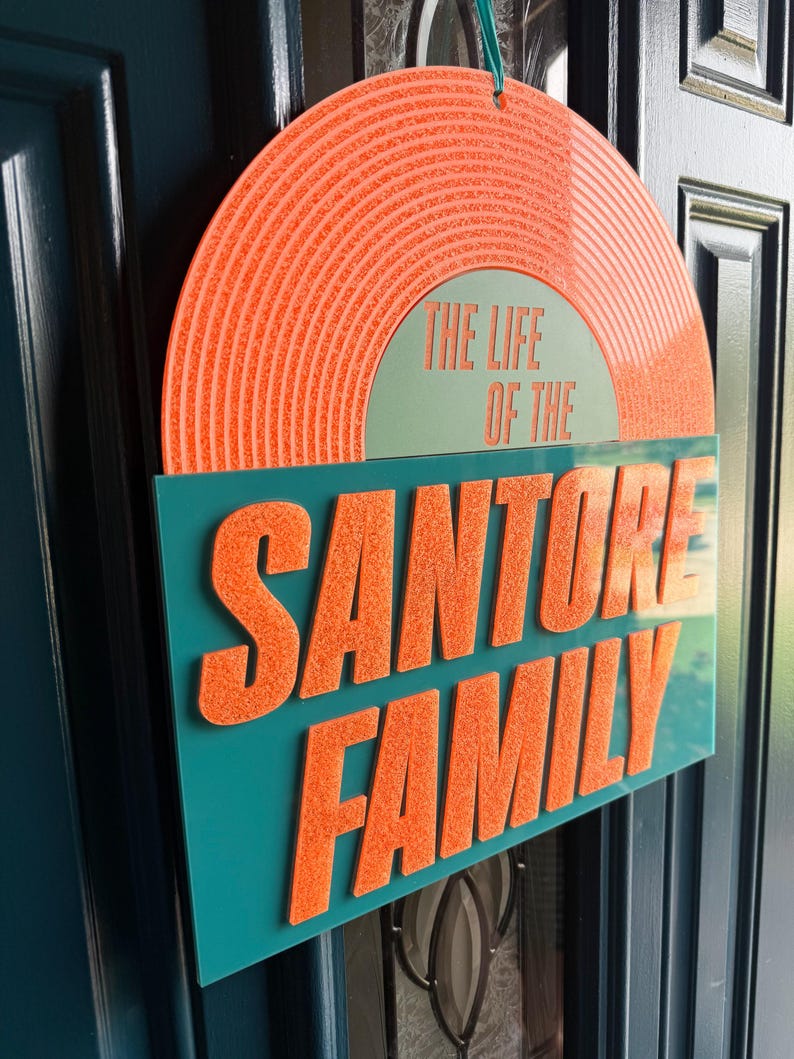 May include: A decorative sign with the text "THE LIFE OF THE SANTORRE FAMILY." The sign features a retro vinyl record design in orange and teal. The text is in a bold, orange font, and the sign is hanging on a teal door.