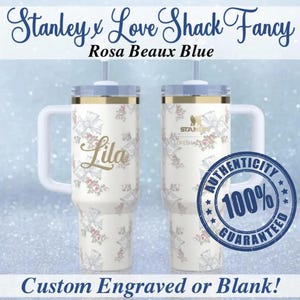 Może przedstawiać: Biały kubek Stanley z kwiatowym wzorem i imieniem "Lila" w złotym skrypcie. Kubek ma uchwyt i pokrywkę. Tekst "Stanley x Love Shack Fancy" i "Rosa Beaux Blue" znajduje się na górze. Tekst "Custom Engraved or Blank!" znajduje się na dole.