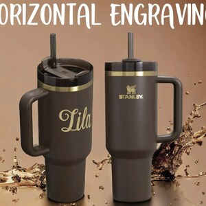Puede incluir: Dos vasos Stanley marrones con asas y detalles dorados. Uno tiene el nombre "Lila" en escritura dorada. El otro presenta el logotipo de Stanley. El texto "HORIZONTAL ENGRAVING" est&aacute; en la parte superior.