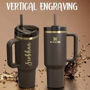 Puede incluir: Dos vasos Stanley marrones con asas y pajitas. Uno est&aacute; grabado con un nombre en escritura dorada. El otro presenta el logotipo de Stanley. El texto "VERTICAL ENGRAVING" est&aacute; en la parte superior.