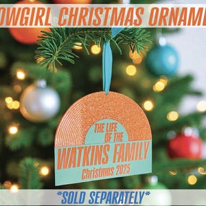 May include: A Christmas ornament shaped like a record, with the text "SHOWGIRL CHRISTMAS ORNAMENT" at the top. The ornament has the text "THE LIFE OF THE WATKINS FAMILY Christmas 2025". It hangs from a blue ribbon on a Christmas tree.