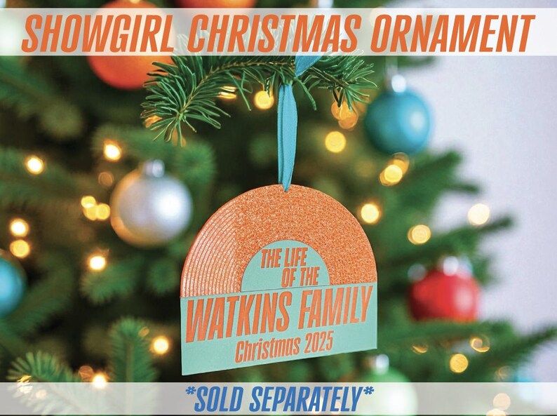 May include: A Christmas ornament shaped like a record, with the text "SHOWGIRL CHRISTMAS ORNAMENT" at the top. The ornament has the text "THE LIFE OF THE WATKINS FAMILY Christmas 2025". It hangs from a blue ribbon on a Christmas tree.