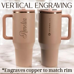 May include: Two insulated tumblers with handles, in a light pink hue, are displayed. One features the name "Amelia" engraved vertically, while the other has "HYDROJUG" printed on it. The rims and handles are a matching copper colour. Text at the top reads "VERTICAL ENGRAVING".