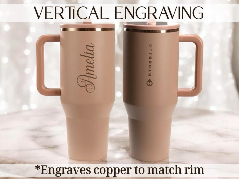 May include: Two insulated tumblers with handles, in a light pink hue, are displayed. One features the name "Amelia" engraved vertically, while the other has "HYDROJUG" printed on it. The rims and handles are a matching copper colour. Text at the top reads "VERTICAL ENGRAVING".