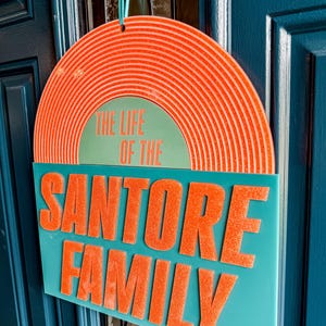 May include: A teal family sign with the text "THE LIFE OF THE SANTORE FAMILY" in orange. The top portion is a half-circle with orange concentric lines. The sign is hanging on a blue door.