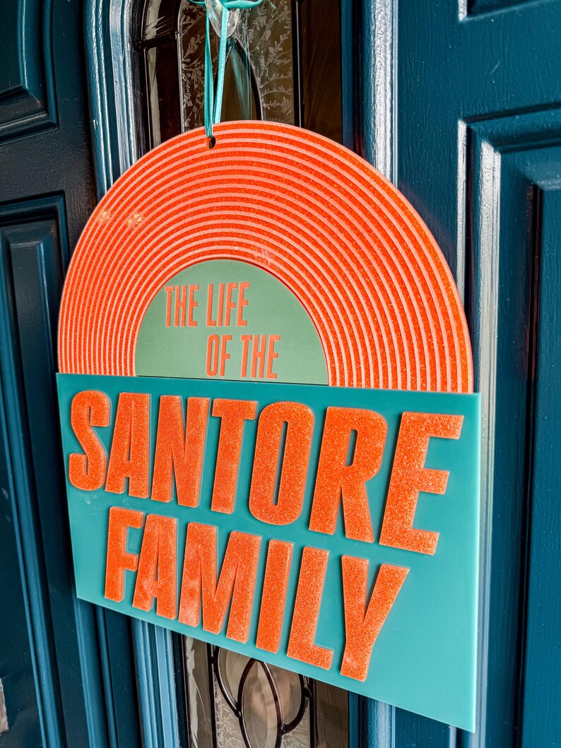 May include: A teal family sign with the text "THE LIFE OF THE SANTORE FAMILY" in orange. The top portion is a half-circle with orange concentric lines. The sign is hanging on a blue door.