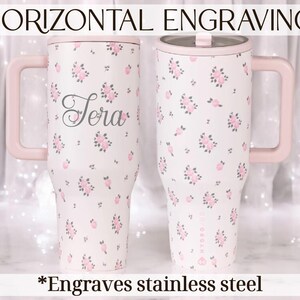May include: Two stainless steel tumblers with pink handles and lids. The tumblers are white with a pink floral pattern and the name "Sera" is engraved on one. The text "Horizontal Engraving" and "*Engraves stainless steel" are also visible.