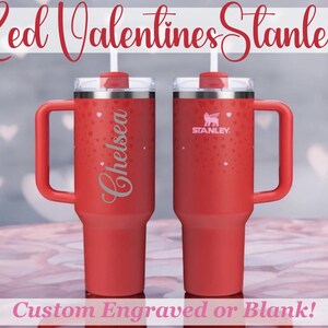May include: Two red Stanley travel mugs with handles and lids. One mug is engraved with the name "Chelsea" and the other has the Stanley logo. Both mugs have a pattern of small hearts.