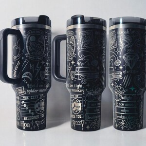 May include: Three black travel mugs with silver etched designs and handles. Each mug features unique illustrations, including text, lightning bolts, and a diamond. The mugs have a black lid and a handle. The text on the mugs includes "Hold on tight, spider monkey" and "Twilight, new moon, eclipse".