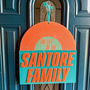 May include: A door sign with the text "THE LIFE OF THE SANTORE FAMILY." The sign has a teal rectangular base with orange, raised lettering. The top is a semi-circle with orange concentric lines and the text "THE LIFE OF THE" in green.
