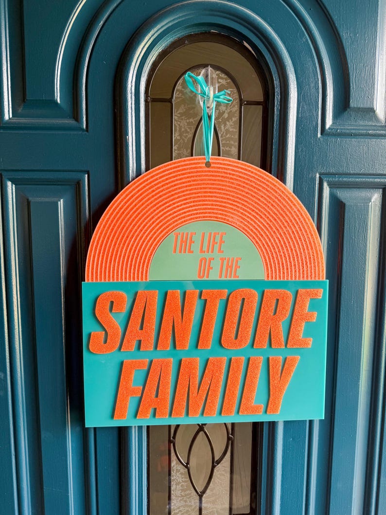 May include: A door sign with the text "THE LIFE OF THE SANTORE FAMILY." The sign has a teal rectangular base with orange, raised lettering. The top is a semi-circle with orange concentric lines and the text "THE LIFE OF THE" in green.