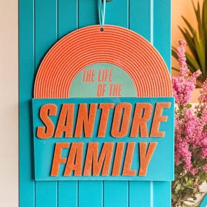 May include: A teal door with a hanging sign. The sign features a retro design with an orange vinyl record shape at the top. Below, a teal rectangle displays the text "THE LIFE OF THE SANTORE FAMILY" in orange, retro-style lettering.