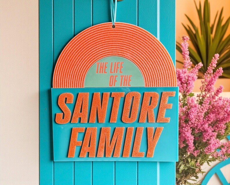May include: A teal door with a hanging sign. The sign features a retro design with an orange vinyl record shape at the top. Below, a teal rectangle displays the text "THE LIFE OF THE SANTORE FAMILY" in orange, retro-style lettering.