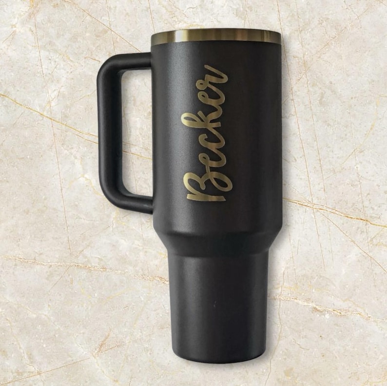 Puede incluir: Un termo de viaje negro con asa y borde dorado. El nombre "Becker" est&aacute; escrito en letras doradas en el lateral. El termo est&aacute; dise&ntilde;ado para mantener las bebidas calientes o fr&iacute;as.