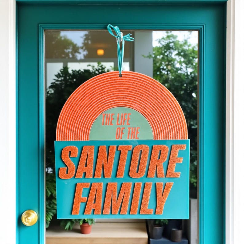May include: A decorative sign with the text "THE LIFE OF THE SANTORE FAMILY." The sign features a retro orange vinyl record design above a teal rectangle with orange lettering. It hangs on a teal door.