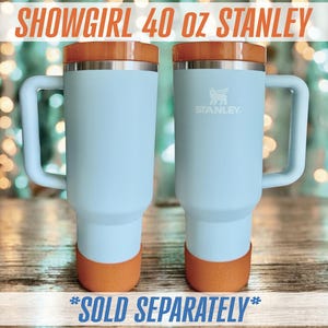 May include: Two light blue Stanley 40 oz tumblers with orange accents on the base and lid. Each tumbler has a handle and the Stanley logo. The text "SHOWGIRL 40 OZ STANLEY" is at the top.