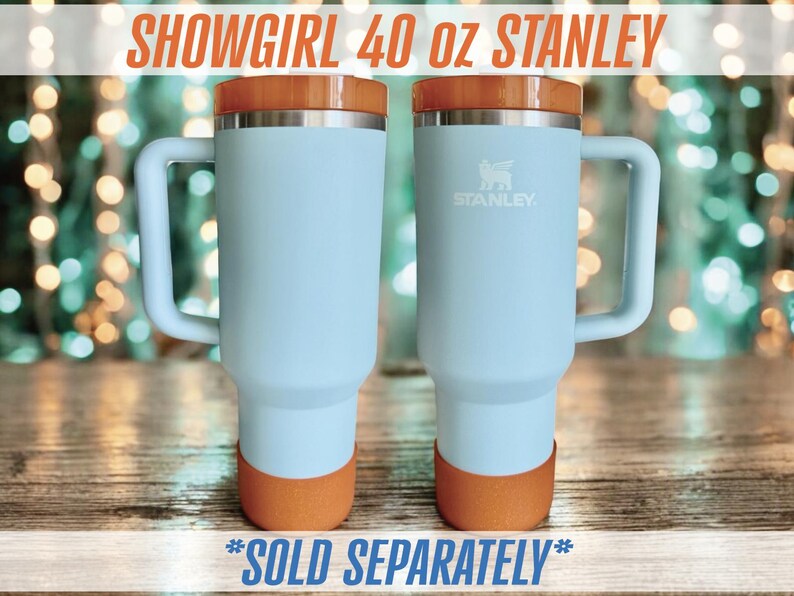 May include: Two light blue Stanley 40 oz tumblers with orange accents on the base and lid. Each tumbler has a handle and the Stanley logo. The text "SHOWGIRL 40 OZ STANLEY" is at the top.