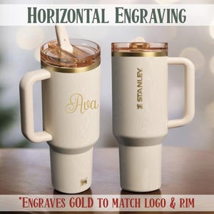 May include: Two cream-colored Stanley tumblers with gold accents and handles. One tumbler is engraved with the name "Ava" in gold script. The other features the Stanley logo. Both have gold rims and lids. The text "HORIZONTAL ENGRAVING" is at the top.