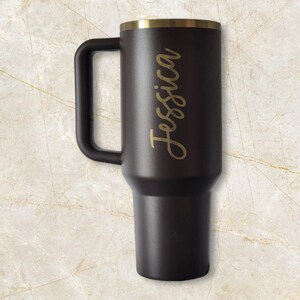 Puede incluir: Un termo de viaje negro con asa y borde dorado. El nombre "Jessica" est&aacute; escrito en letras doradas en el lateral. El termo est&aacute; dise&ntilde;ado para mantener las bebidas calientes o fr&iacute;as.